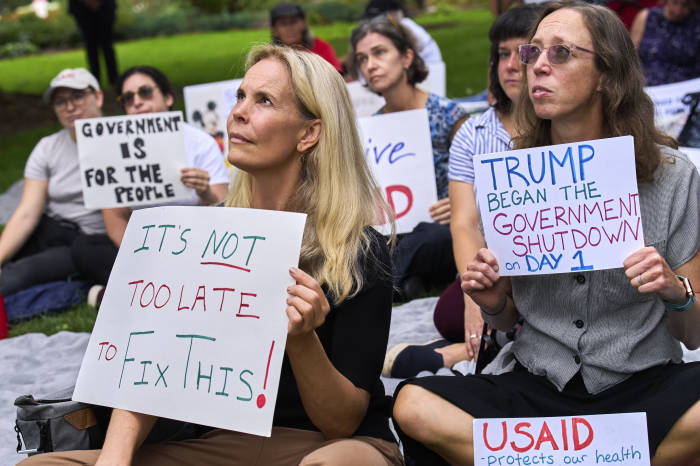 Demonstrators hold signs about the government shutdown, one reading “It’s Not Too Late to Fix This!” and the other “Trump Began the Government Shutdown on Day 1.”