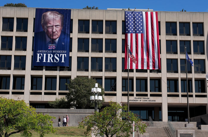 A large banner with an image of Donald Trump that reads "American Workers First" hangs on the Department of Labor building, next to a large American flag.