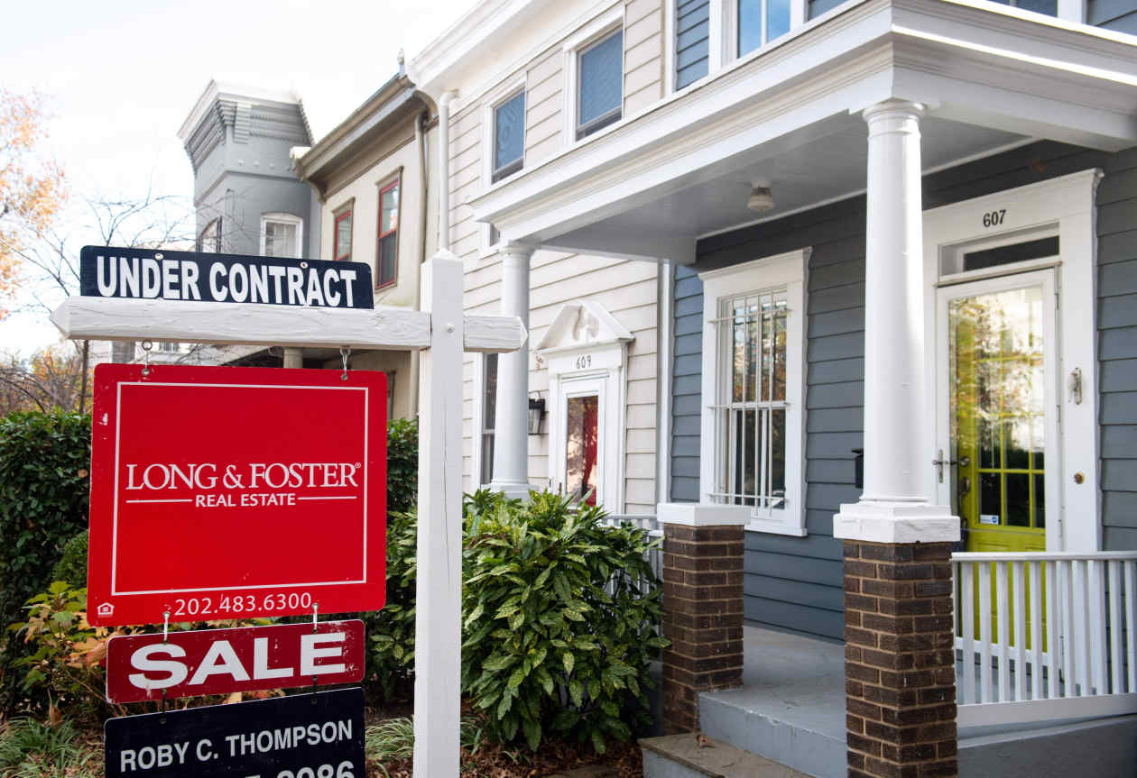 A "Long & Foster Real Estate" for sale sign, with an "UNDER CONTRACT" banner, stands in front of a blue townhouse with a white porch.