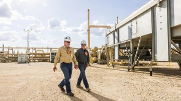 Two BPX Energy workers walk on a dirt path in an industrial setting.