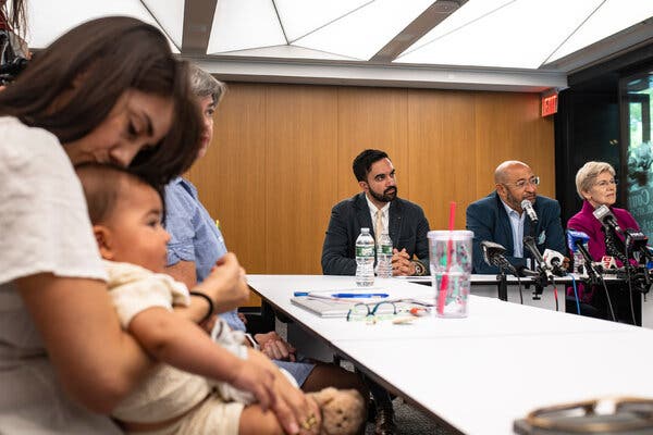 Mr. Mamdani sits during a news conference, with Senator Elizabeth Warren to his far left, and a young child in the foreground, held in the lap of a woman.