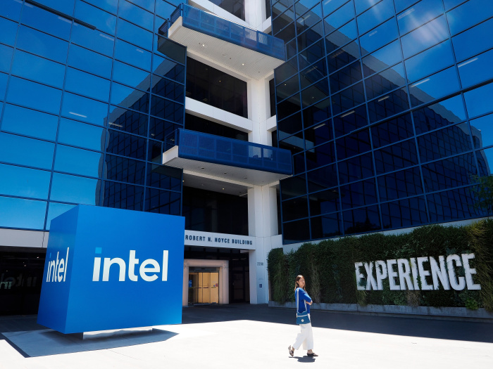 The Intel Corporate Headquarters in Santa Clara, California, with a large blue Intel logo and a woman walking in front of the building.