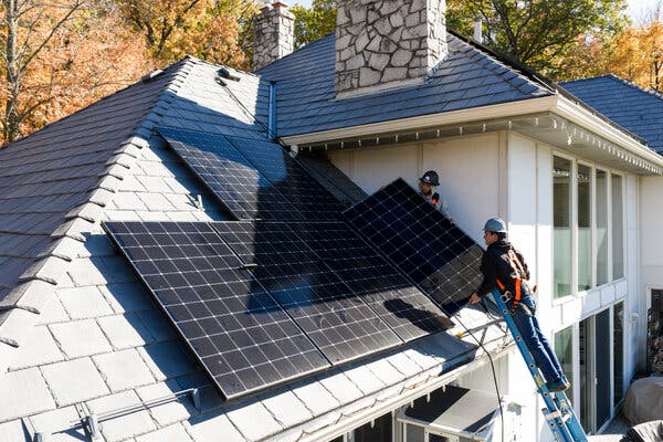 Two workers, one of them on a ladder, place a solar panel on the roof of a house. 