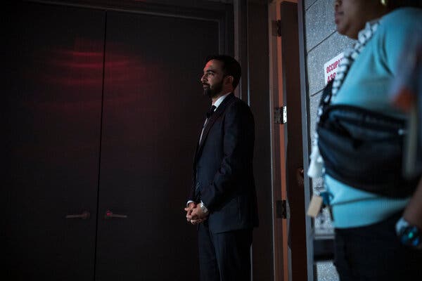 Mr. Mamdani stands outside two wooden doors, his hands clasped in front of him.