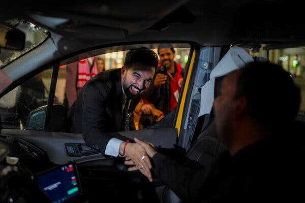 Mr. Mamdani broadly smiles as he reaches into a taxi to shake the hand of a driver.