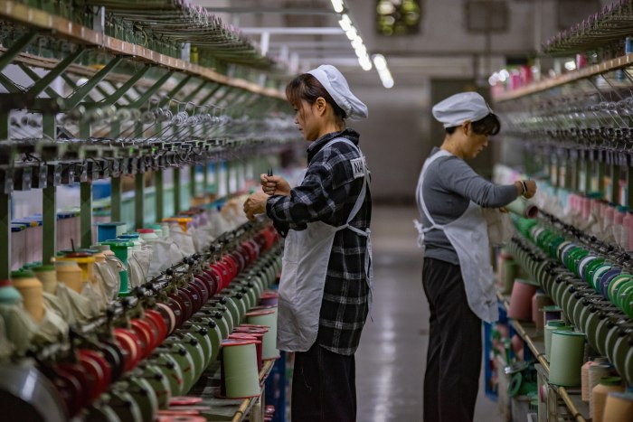 Two silk factory workers in white aprons work along a production line with colorful thread spools in Fuyang, China.