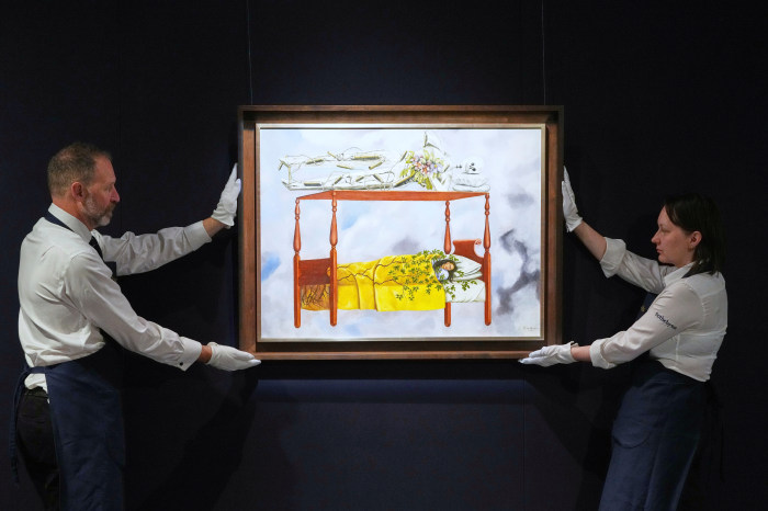 Two art handlers in white gloves hold up Frida Kahlo's painting titled “El sueño (La cama)” or (The Dream (The Bed)).