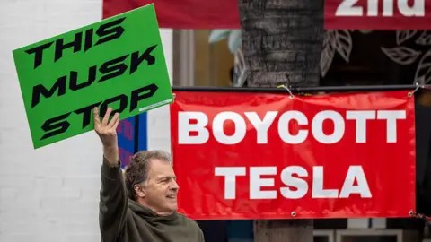 Getty Images A man holds a placard saying "This Musk Stop" in front of a banner reading "Boycott Tesla"