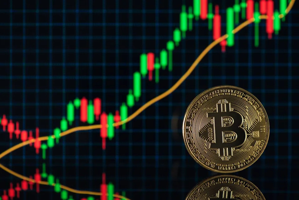 A golden Bitcoin coin stands upright on a reflective dark surface in the right foreground. Behind it, a blurred digital display shows a financial candlestick chart with numerous red and green bars, indicating market movement, alongside a prominent curving yellow trend line against a dark grid background.