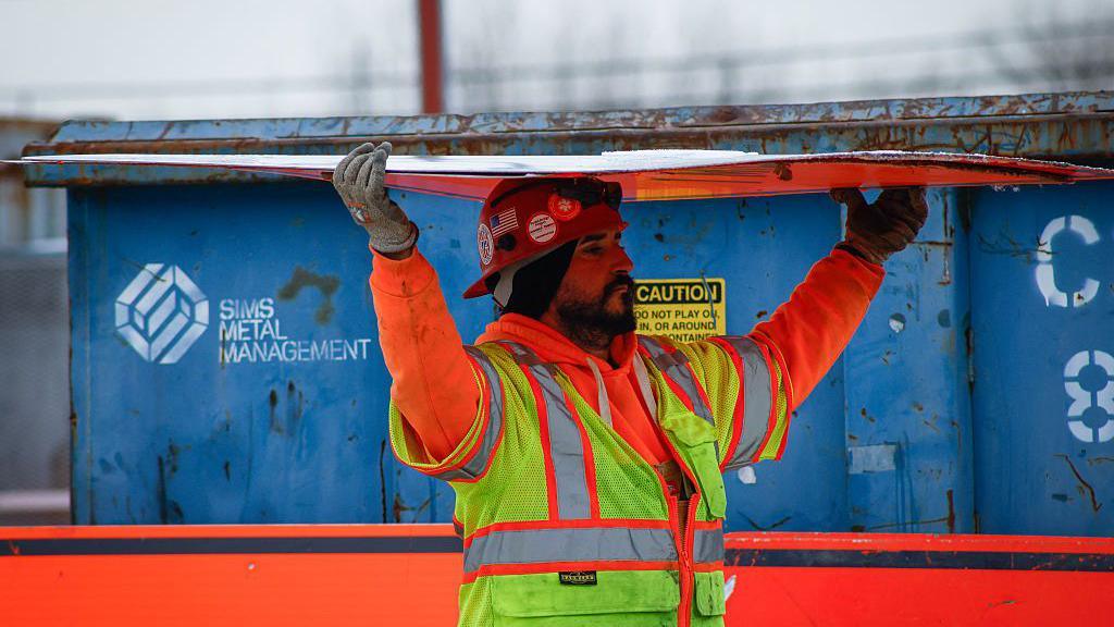 A bearded construction worker wearing an orange sweatshirt and yellow safety vest carries a sheet of construction material over his his head.  In the background there is a a blue container. The picture was taken at the site of the Gateway Tunnel construction project as seen from North Bergen, New Jersey, on February 10, 2026.