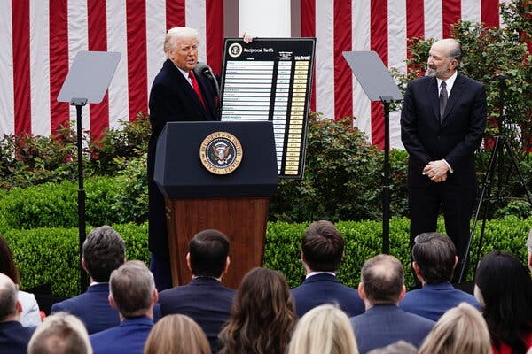 President Trump, standing behind a lectern, holds up a cardboard placard that lists countries and tariffs. Behind him is the White House, with big American flags hanging down the walls.