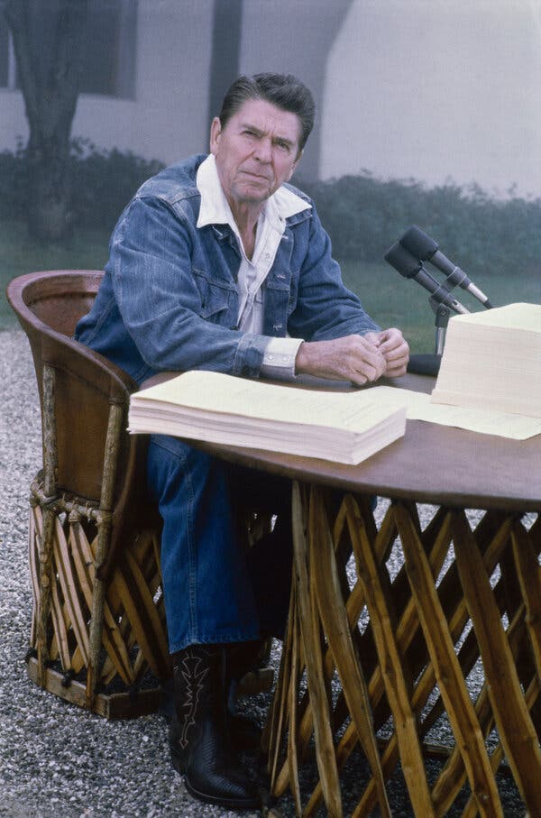 Ronald Reagan, wearing a jean jacket over a white shirt, sits at a desk in the driveway near a house. On the table are microphones and large piles of paper.