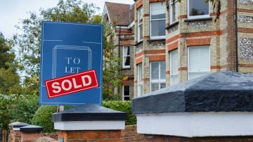 "To Let" sign with "Sold" sticker over it, in front of a brick building.