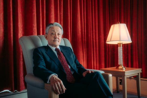 A portrait of Hiromi Yamaji, in a suit and sitting in an upholstered chair next to a table and lamp, looking at the camera.