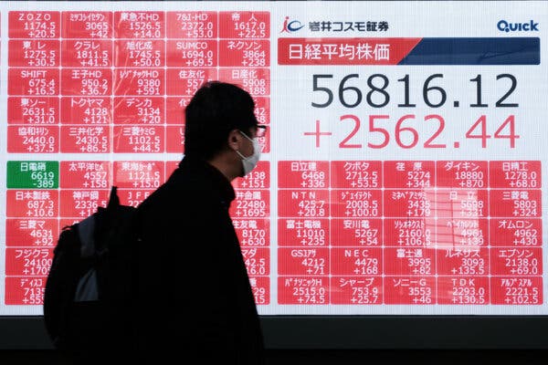 A person in a dark jacket and face mask walks by a digital board. White numbers "56816.12" and red "+2562.44" appear with other stock data.