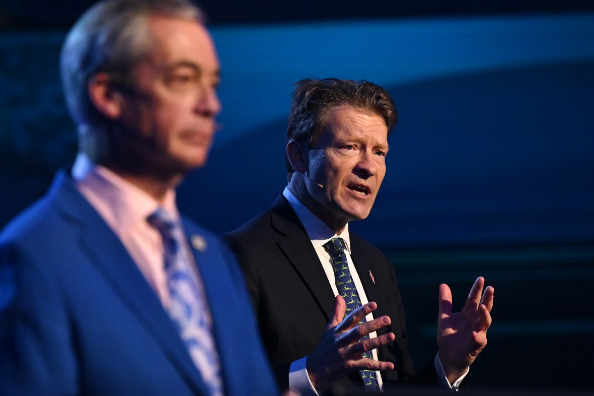 Richard Tice speaking at a podium while Nigel Farage is blurred in the background.