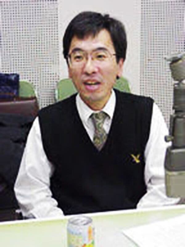 A photo of Mr. Kubota taken when he was a young dealer, sitting a desk, wearing a sweater vest and tie, talking to someone. 