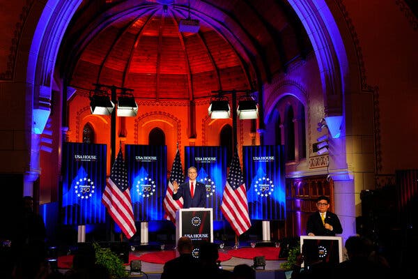 Scott Bessent speaking from behind a lectern with American flags and blue USA House signs behind him.