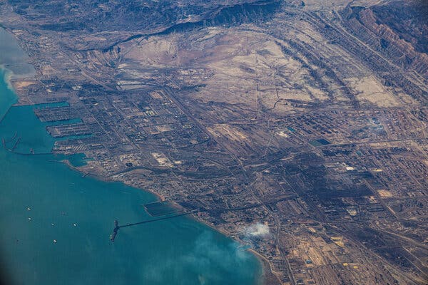 Aerial view of a blue bay with a large port and coastal development. Dry, rugged land extends toward distant mountains.