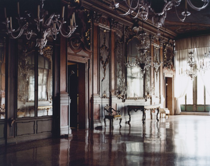 An ornate room with chandeliers, mirrored walls, and dark wood paneling, featuring a polished floor reflecting the light.