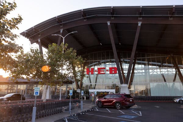 A couple cars are parked outside a glass-walled store with the large red letters spelling H-E-B on the front.