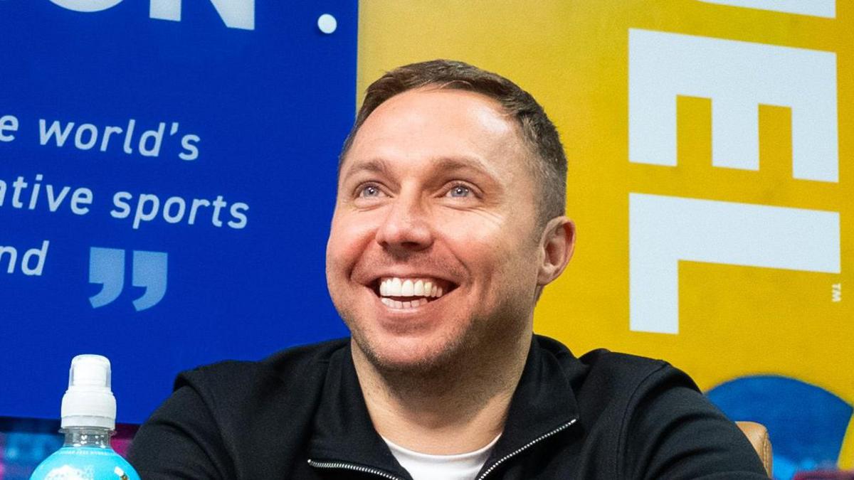 Tom Ryder, who is smiling, is sitting a desk in front of a range of Applied Nutrition products. There are a number of Applied Nutrition advertisements in the background. 