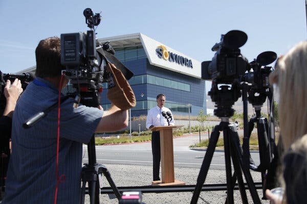 Mitt Romney, wearing a white shirt and dark pants, stands at a lectern. Behind him is an office building with the name Solyndra on the side.