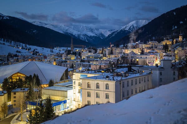 A snow-covered town at dusk, buildings illuminated against dark mountains. A large modern structure with an angular roof is in the foreground.