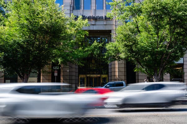 The front of a stone building with the words The Washington Post over the entrance. Blurred cars going by on a street are seen in the foreground.