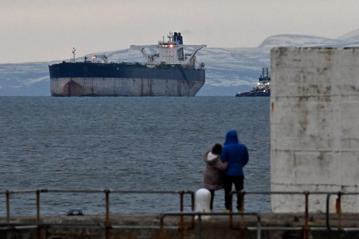 Two people on a pier look out at an oil tanker and a smaller vessel in the water.