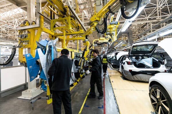 A factory assembly line with people working on cars. Overhead yellow machinery transports car components for white cars that await completion.
