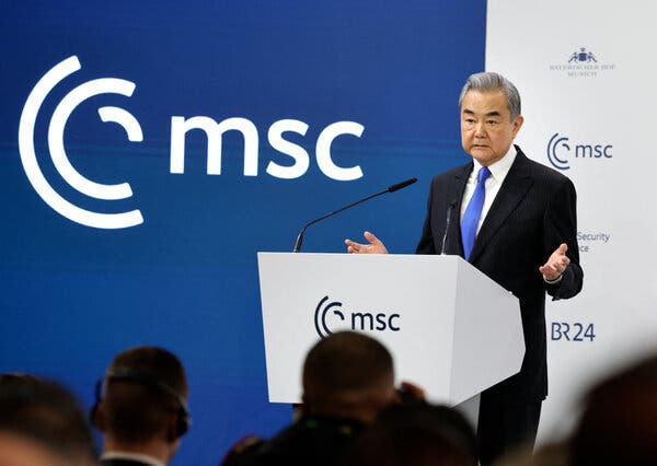 Wang Yi, wearing a dark suit, white shirt and blue tie, stands at a white lectern, gesturing with open hands. 
