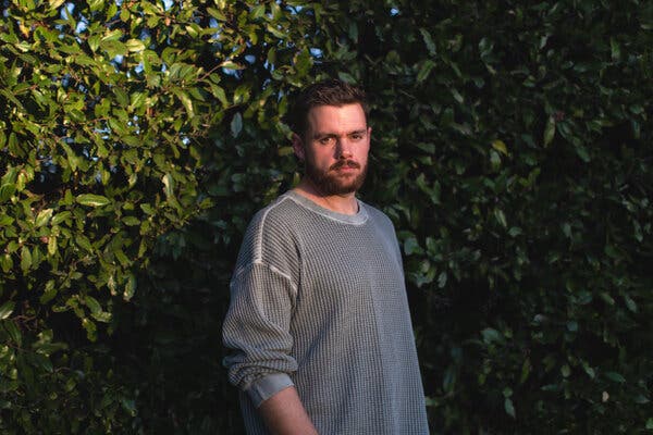A man in a gray shirt stands in front of trees.