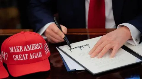 Getty Images US President Donald Trump signs an executive order in the Oval Office at the White House in Washington, DC on 23 April 2025.  A red baseball hat embroidered with "Make America Great Again" in white thread sits to the left of the document, which shows Trump's distinctive signature