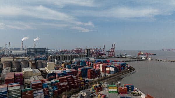 A vast industrial port with many colorful containers stacked high. Smokestacks emit white smoke, with wind turbines also visible.