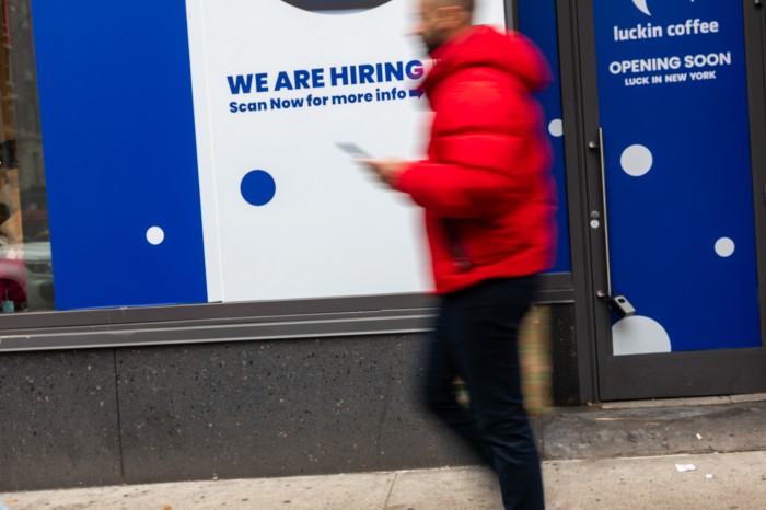 A "We Are Hiring" sign is displayed in a business's window next to a blurred figure walking past.